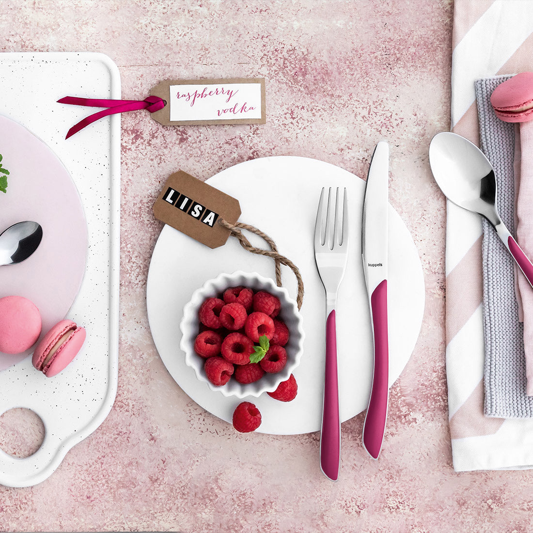 Ein gedeckter Tisch mit rosafarbenem Besteck, einer Schüssel mit Himbeeren, rosafarbenen Macarons, einem Löffel, einem Messer, einer Gabel und zwei Namensschildern mit der Aufschrift Liisa und Himbeerwodka auf einem rosafarbenen Hintergrund.