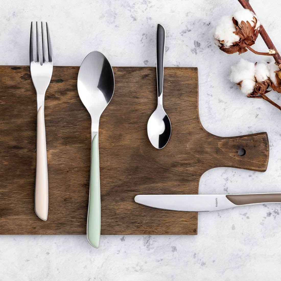 Das 24-teilige PRISMA Besteckset in Salbeigrün von Kuppels mit Gabeln, Esslöffeln und Teelöffeln mit bunten Griffen ist elegant auf einem Holzbrett mit einem Messer mit beigem Griff und Baumwollzweigen auf Marmor angerichtet.