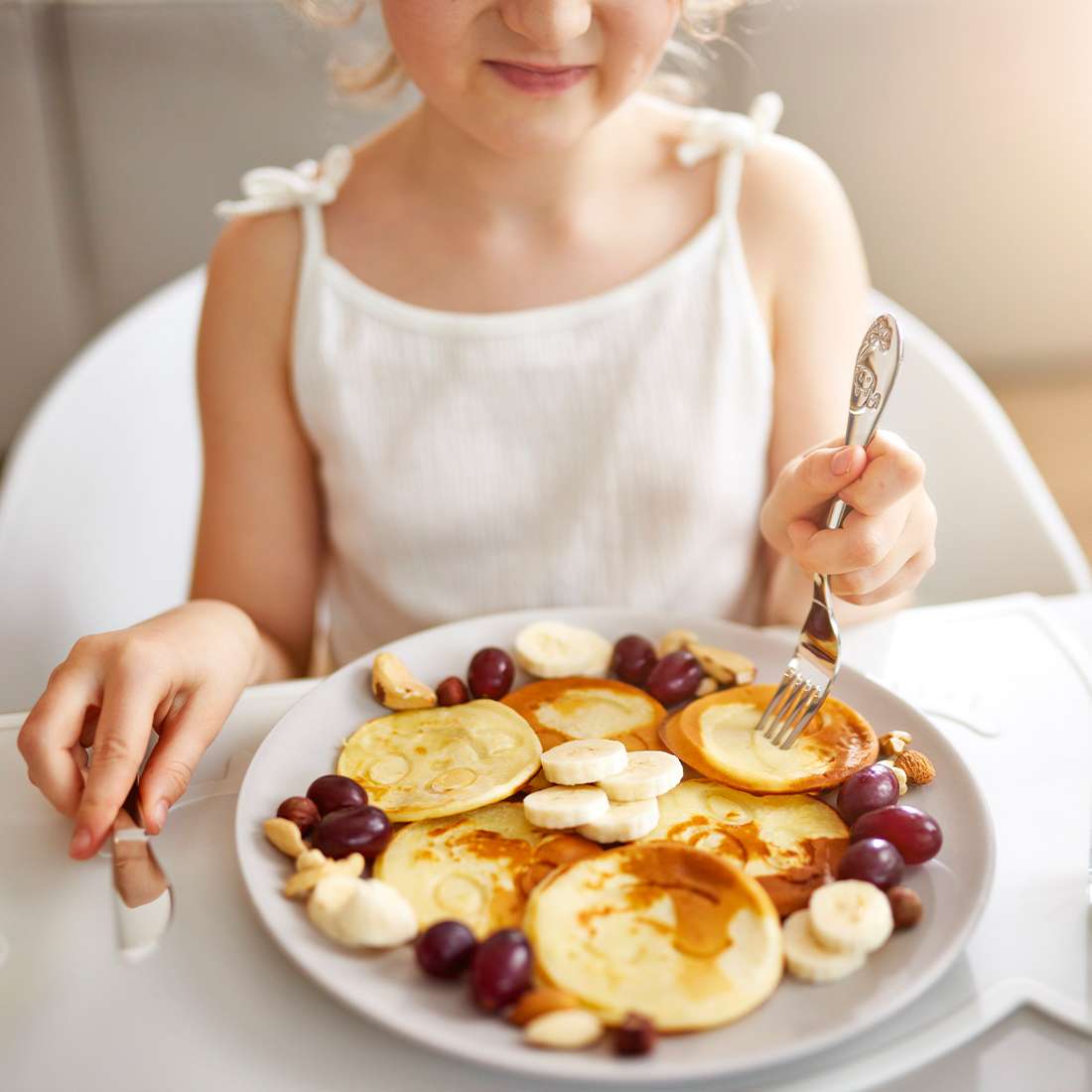 Ein Kind in einem weißen Tanktop lächelt an einem Tisch und hält das 4-teilige Kuppels BAUERNHOFFREUNDE Kinderbesteck-Set mit niedlichen Bauernhoftiere-Motiven vor einem mit Pfannkuchen, Bananen, Weintrauben und Nüssen dekorierten Teller.