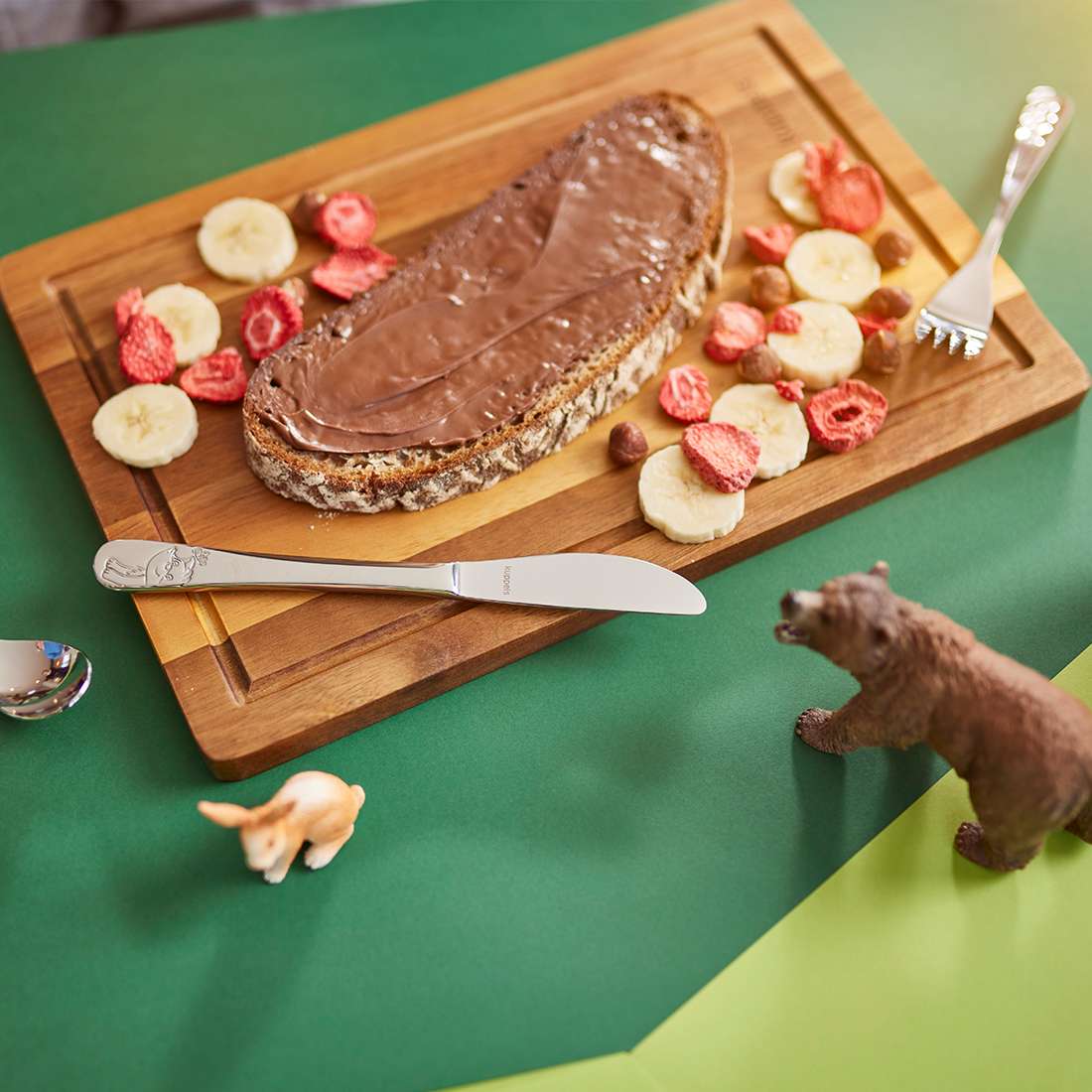 Eine Scheibe Mehrkornbrot mit Schokoaufstrich liegt auf einem Holzbrett, umgeben von Bananenscheiben und getrockneten Erdbeeren. Daneben stehen das 4-teilige Kuppels WALDFREUNDE Kinderbesteck-Set und verspielte Bären- und Schweinefiguren auf dem grünen Tisch.