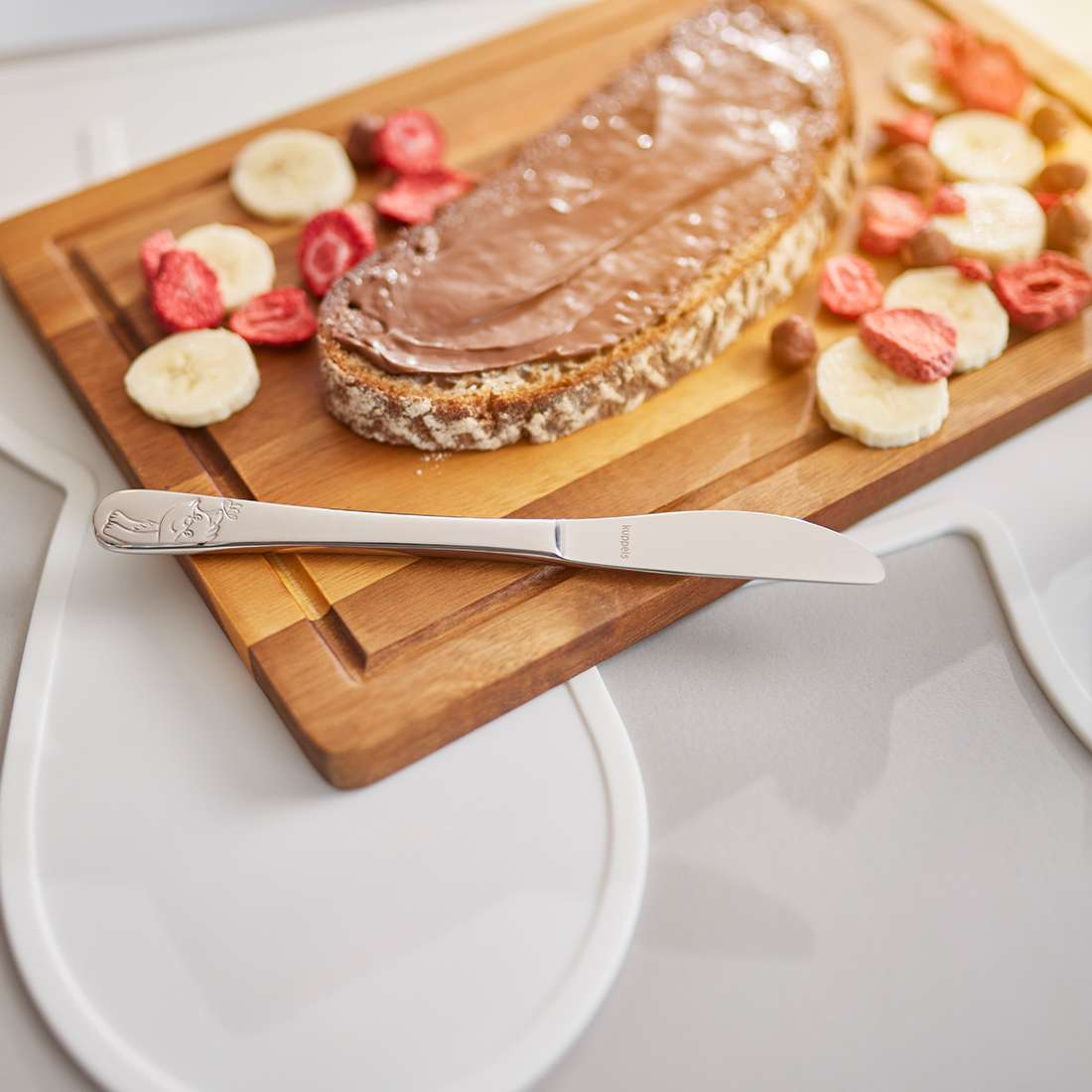 Auf einem Holzbrett liegt eine Brotscheibe mit Schokoladenaufstrich, umgeben von Bananenscheiben und getrockneten Erdbeeren, davor ein silbernes Kuppels WALDFREUNDE Kinderbesteck-Set 4-teilig Messer (Edelstahl 18/10).