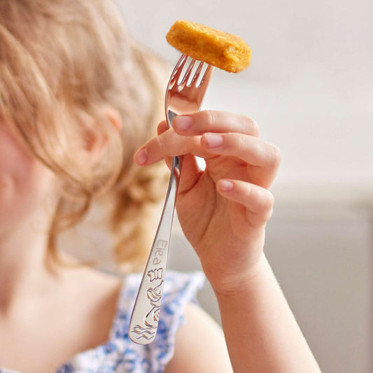 Ein Kind hält eine Gabel aus dem 4-teiligen DUCK-Kinderbesteck-Set in der Hand, auf deren Zinken ein Chicken Nugget aufgespießt ist; auf dem Griff sind ein Name und Entenmotive eingraviert.