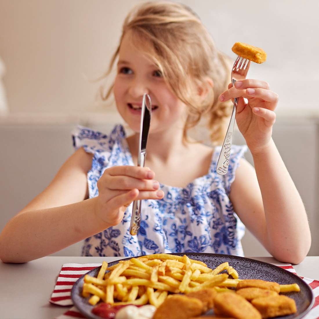 Ein junges Mädchen in einem blau-weißen Kleid lächelt und hält das Kuppels DUCK Kinderbesteck-Set 4-teilig mit Chicken Nuggets und Messer in der Hand. Vor ihr steht ein Teller mit Pommes Frites, Chicken Nuggets und Dip-Saucen.