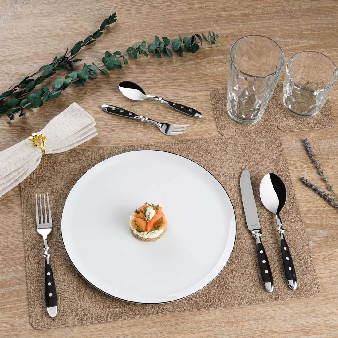 Ein Esstisch im Landhausstil: ein weißer Teller mit einer Vorspeise, Kuppels COUNTRY Besteckset 24-teilig schwarz, zwei leere Gläser, eine beigefarbene Serviette mit Goldring, Zweige als Dekoration, alles auf einem Holztisch mit braunem Tischset.
