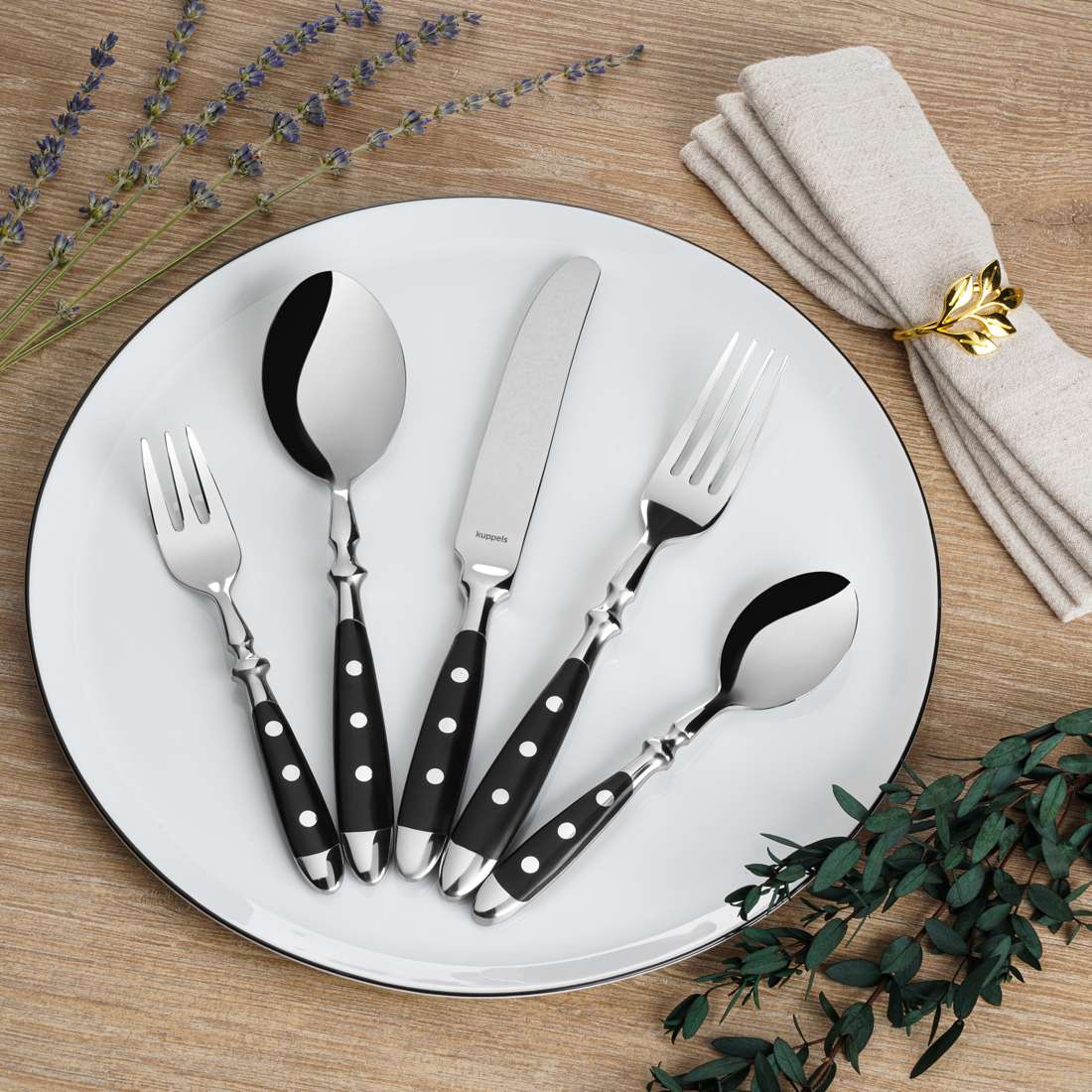 Das Kuppels COUNTRY Besteckset 24-teilig schwarz zeichnet sich durch elegante schwarze Griffe aus, die auf einem weißen Teller mit Lavendel, Grünzeug und einer Serviette wunderschön zur Geltung kommen - ideal für ein Ambiente im Landhausstil.