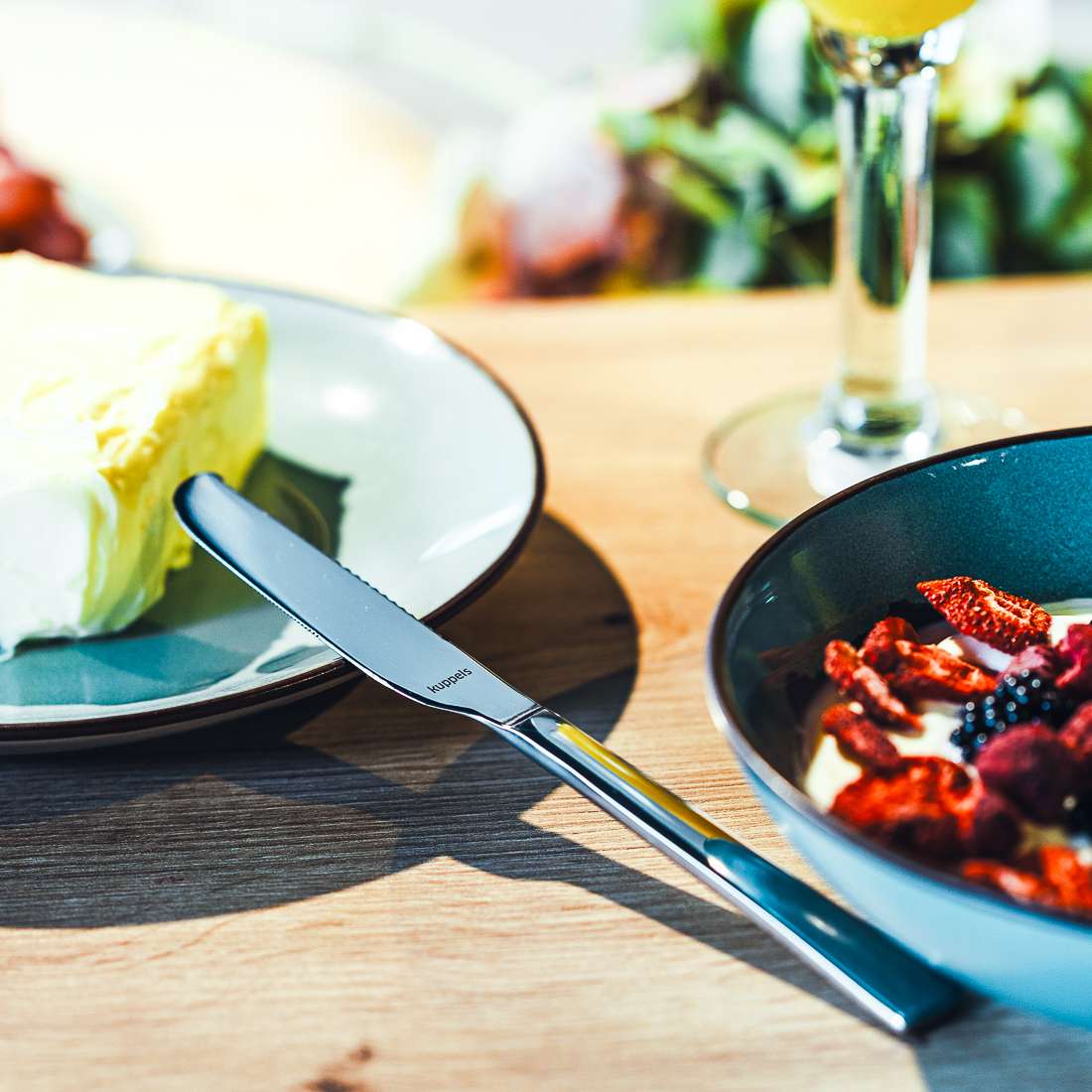 Eine Nahaufnahme zeigt einen Kuchenteller, ein Kuppels SELECTION Butterstreicher Set 2-teilig mit gezackter Klinge am Rand, eine Schale Joghurt mit Beeren und Trockenfrüchten und im Hintergrund ein Glas Weißwein.