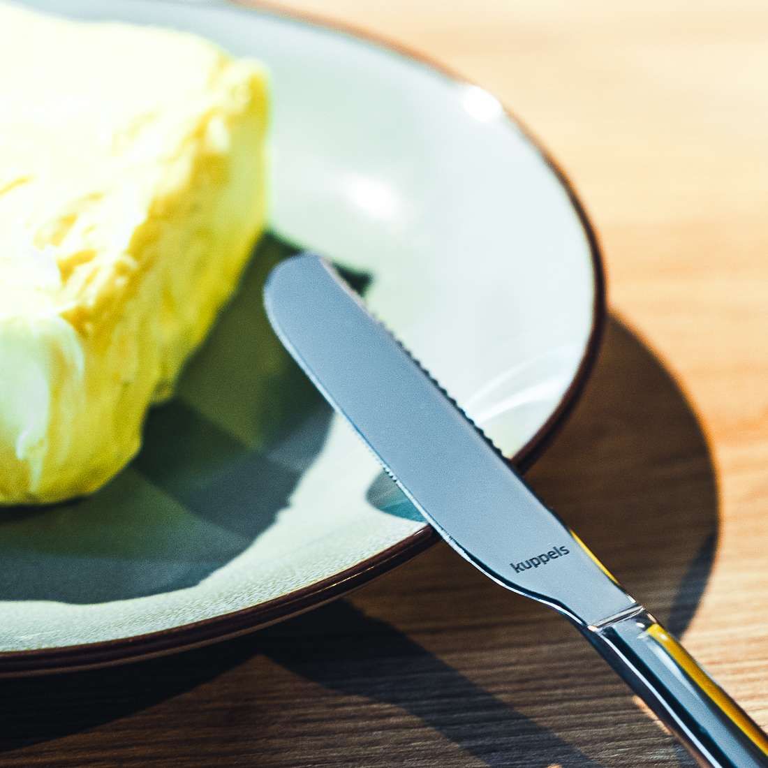Eine Nahaufnahme des SELECTION Butterstreicher-Sets 2-teilig von Kuppels zeigt ein Messer mit der Aufschrift „Kuppels“ am Griff, das neben Butter auf einem Teller liegt. Seine gezackte Klinge sorgt für ein gleichmäßiges Streichen in dieser warmen, einladenden Szene.