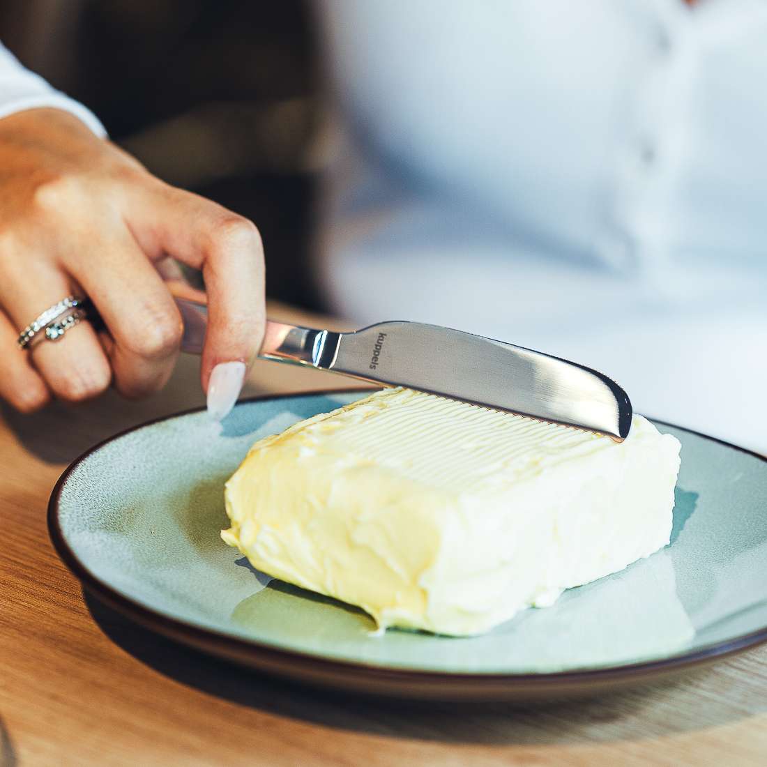 Eine manikürte Hand mit Ring streicht mit dem Kuppels SELECTION Butterstreicher Set 2-teilig Butter von einem rechteckigen Block auf einen grünen Teller, vor einem sanft verschwommenen Hintergrund.