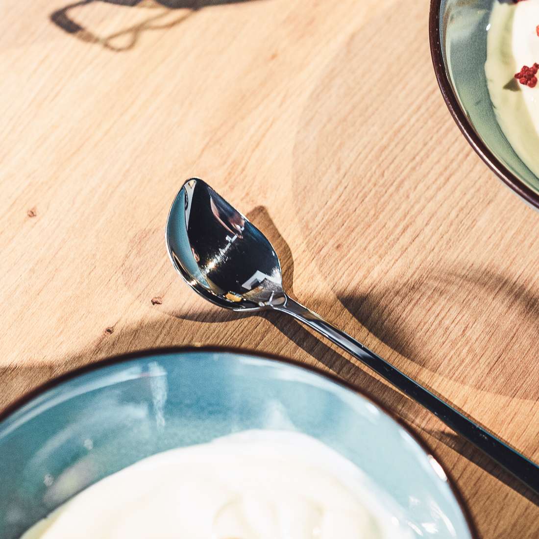 Ein Kuppels SELECTION Joghurtlöffel Set 2-teilig aus Edelstahl 18/10 liegt auf einem Holztisch neben zwei blau umrandeten Schalen mit cremigem Joghurt, von denen eine im Vordergrund und eine im Hintergrund zu sehen ist.
