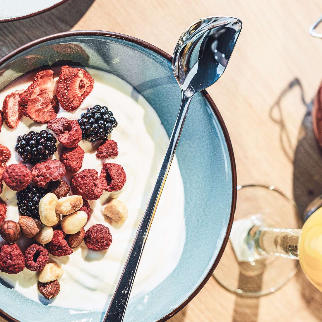 Auf dem Tisch steht eine Schüssel Joghurt mit getrockneten Erdbeeren, Himbeeren, Brombeeren und Haselnüssen. Darin befindet sich ein 2-teiliger Joghurtlöffel-Set von Kuppels SELECTION. Daneben steht ein Glas Orangensaft.