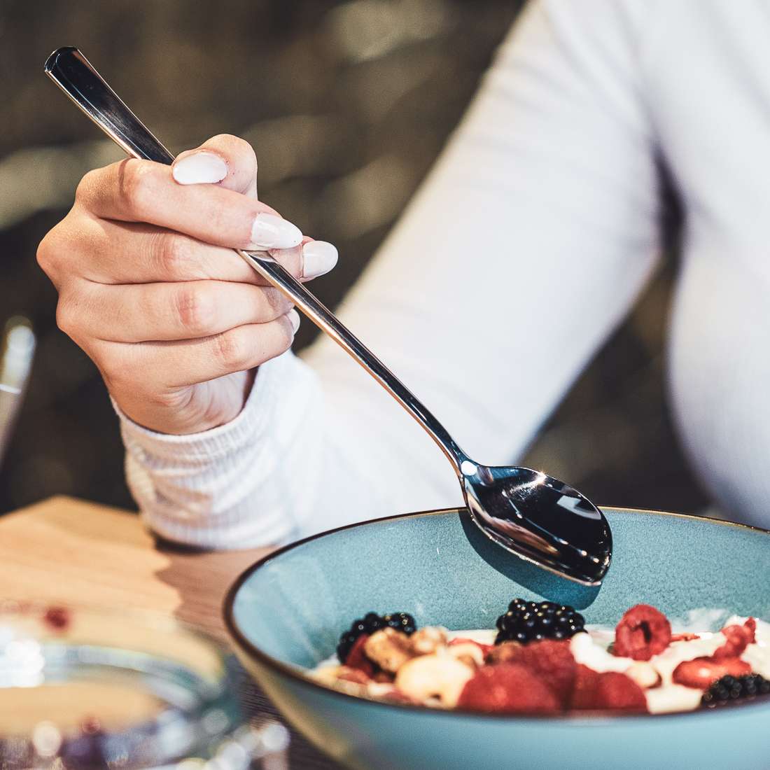 Jemand hält einen Kuppels SELECTION Joghurtlöffel aus dem 2er-Set über eine Schüssel Joghurt mit Himbeeren, Brombeeren und Bananenscheiben und trägt ein weißes Langarmshirt.