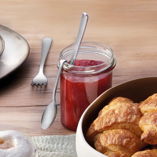 Ein Glas rote Marmelade, in dem ein Marmeladenlöffel von Kuppels SELECTION steckt, steht auf einem Holztisch neben einem Teller mit Croissants und einer Tasse schaumigem Kaffee. Im Hintergrund vervollständigt eine Gabel diese einladende Frühstücksszene.
