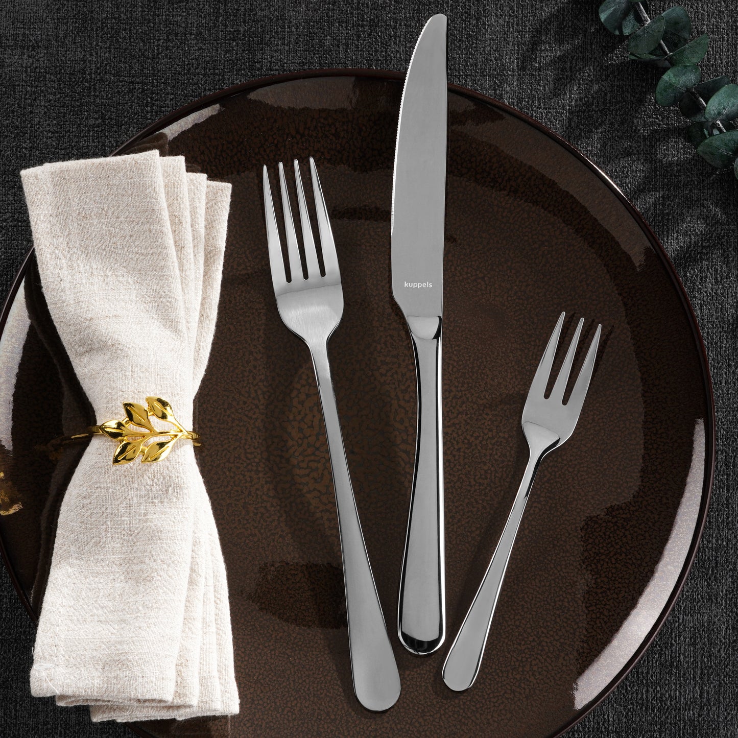 Auf einem braunen Teller liegen eine Gabel, ein Messer und eine Salatgabel aus dem Kuppels DELIA Besteckset 30-teilig (Edelstahl 18/0). Daneben liegt eine cremefarbene Serviette mit einem Ring aus Blattgold, alles auf einer dunkel strukturierten Tischdecke.