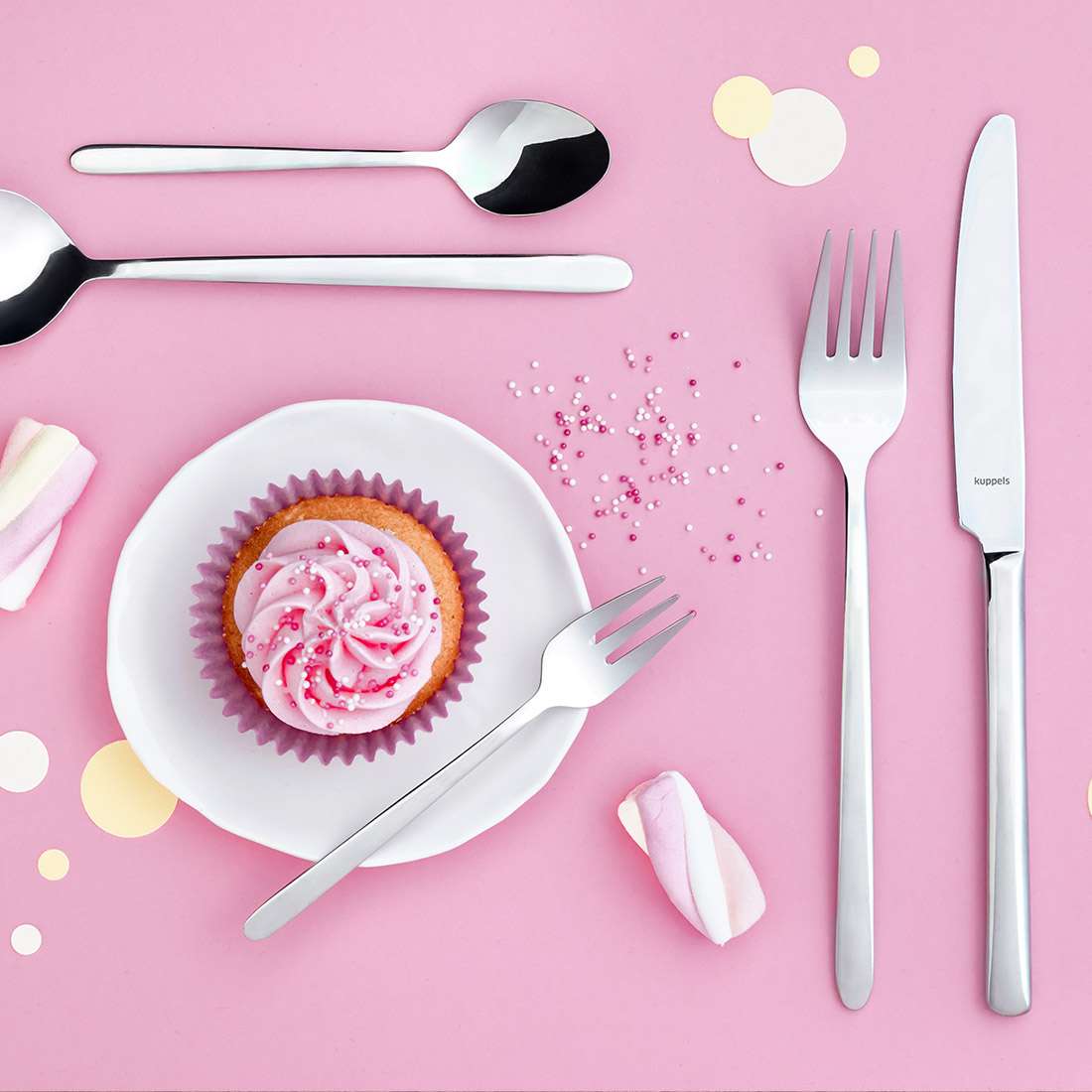 Ein Cupcake mit rosa Zuckerguss auf einem weißen Teller, umgeben vom Kuppels CUTE Besteckset 30-teilig, Marshmallows und Streuseln, arrangiert auf einem pastellrosa Hintergrund mit verstreutem Papierkonfetti.