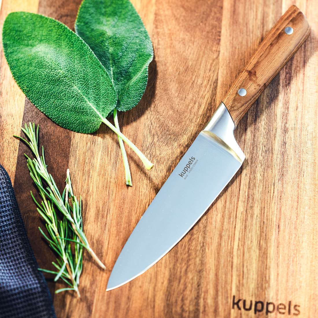Ein Kuppels AVID Kochmesser 6" mit Eschenholzgriff liegt auf einem Holzschneidebrett, beides mit dem Kuppels-Logo gekennzeichnet, neben zwei Salbeiblättern und einem Rosmarinzweig. Die Klinge ist aus 50CrMoV15-Stahl gefertigt.