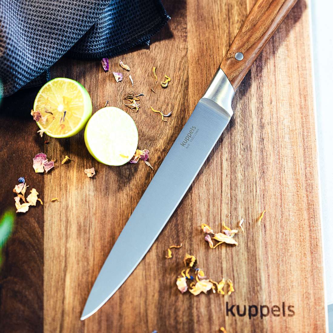 Ein AVID Tranchiermesser von Kuppels mit Eschenholzgriff liegt neben einer halbierten Limette und getrockneten Blütenblättern auf einem Holzschneidebrett. Auf der Ecke des Bretts ist „Kuppels“ aufgedruckt.