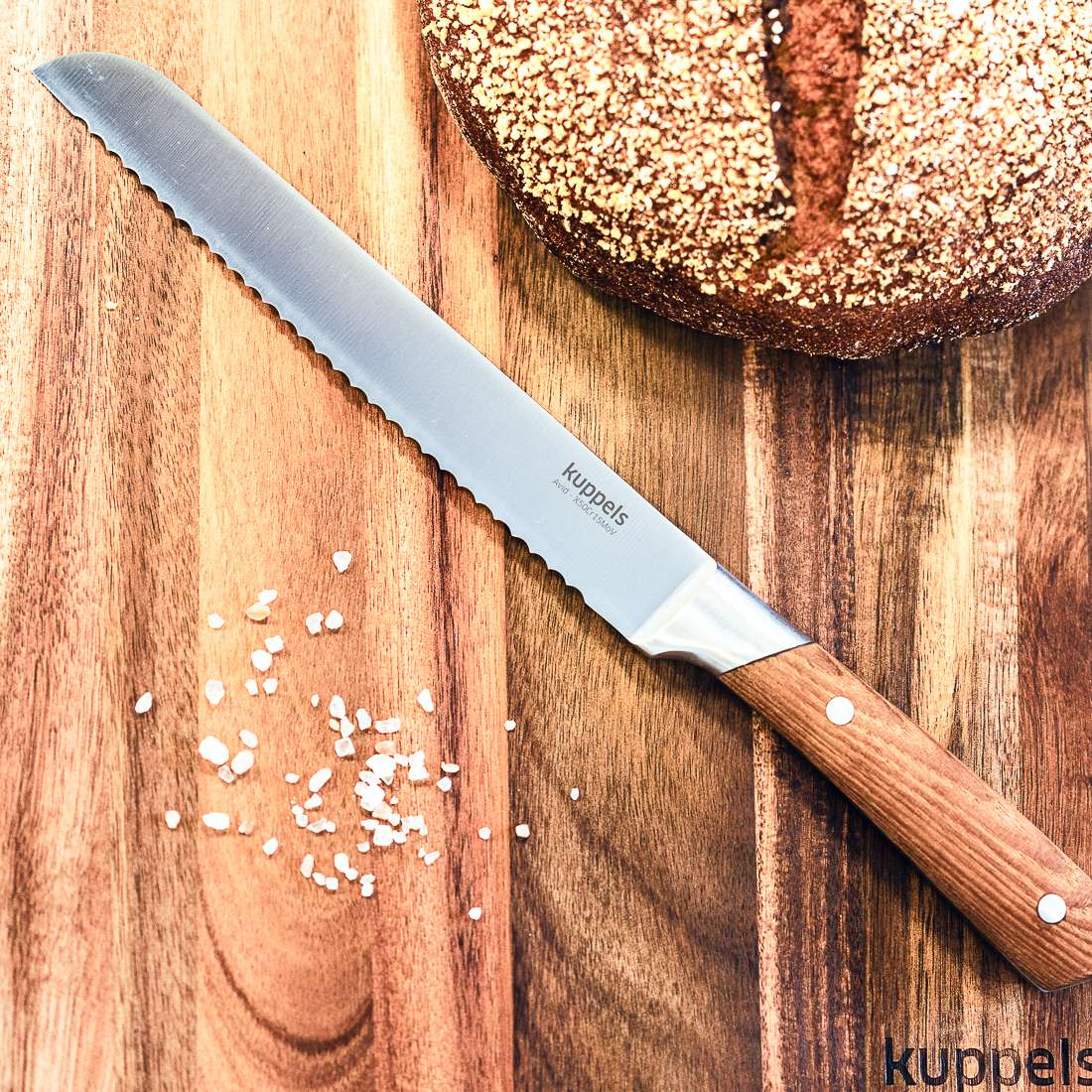 Das AVID Brotmesser von Kuppels mit Wellenschliffklinge und Eschenholzgriff liegt neben einem runden Laib braunen Brots auf einem Holzbrett, umgeben von grobem Salz. Unten rechts ist das Wortkuppels zu sehen.