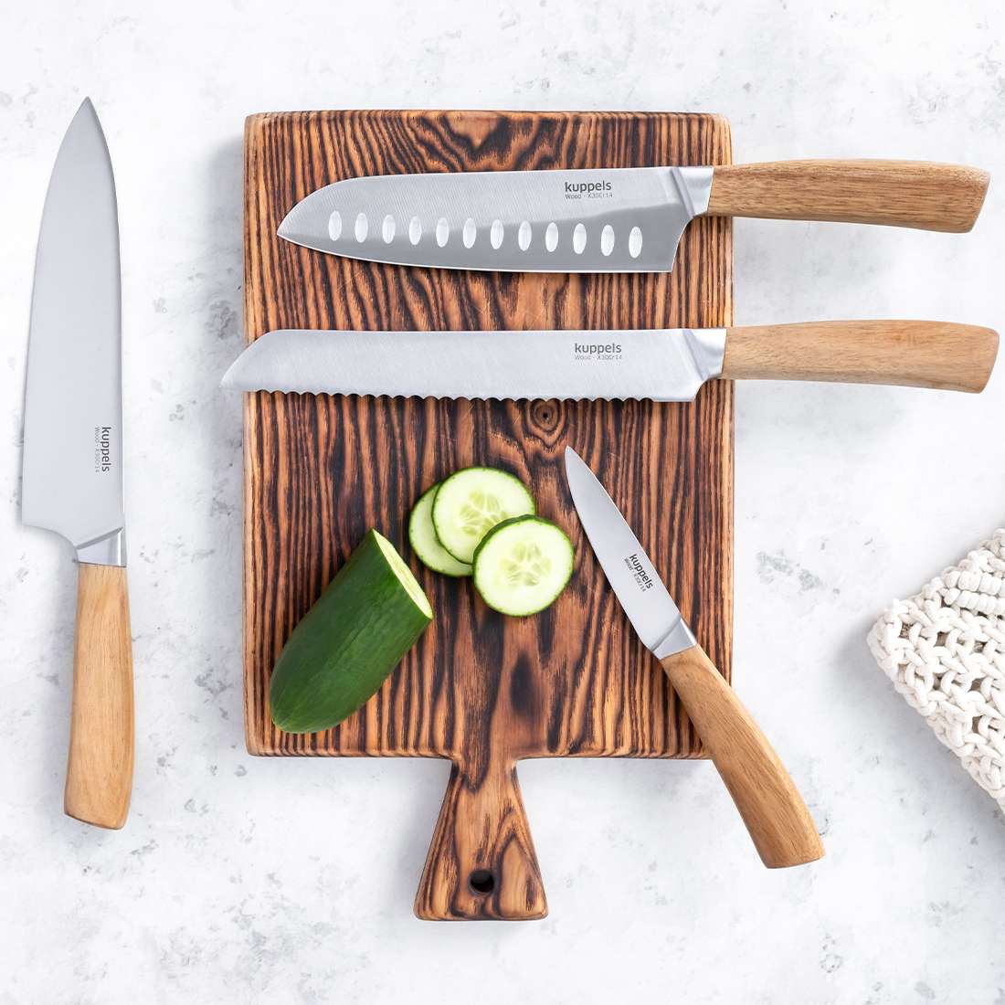 Drei Kuppels WOOD Brotmesser mit Akazienholzgriff und eine angeschnittene Gurke liegen auf einem Holzschneidebrett, ein Messer liegt auf einer hellen Fläche.