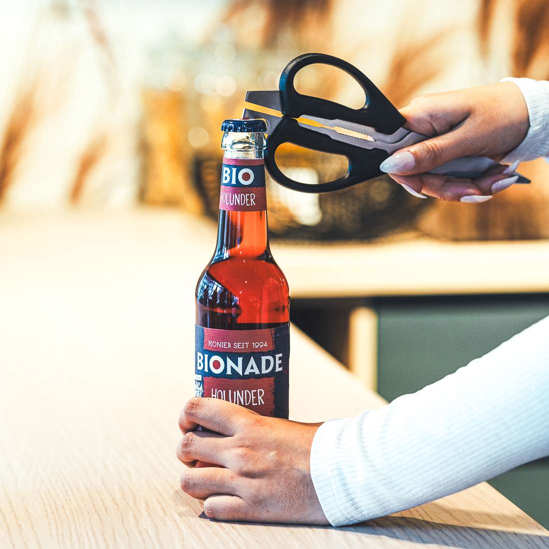 Eine Person öffnet mit einer Kuppels SELECTION Allzweckschere eine Flasche Bionade Holunder und hält die Flasche dabei mit einer Hand auf einer hellen Holzunterlage.