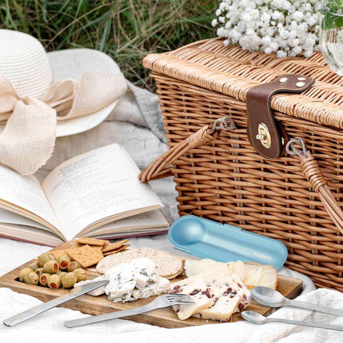 Eine Picknickszene mit Weidenkorb, aufgeschlagenem Buch, Snacks, Sonnenhut und dem Kuppels 2GO Besteckset 4-teilig Mint im kompakten blauen Etui auf einer weißen Outdoordecke.