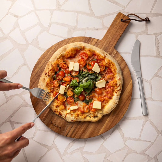 Eine Person schneidet mit einer Gabel und dem 2-teiligen SELECTION-Pizzamesser-Set von Kuppels eine Pizza mit Kirschtomaten, Basilikum und Käsewürfeln auf einem Holzbrett, während daneben ein Messer auf einem Tisch mit Mosaikmuster liegt.