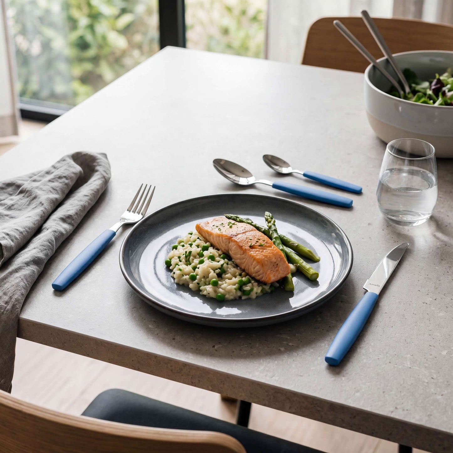 Auf einem Esstisch, der mit einem 16-teiligen hellblauen Amefa VALOA-Besteckset, einem Glas Wasser, einer grauen Serviette und einer Salatschüssel gedeckt ist, steht ein Teller mit gegartem Lachs, Spargel und Risotto in der Nähe eines Fensters.