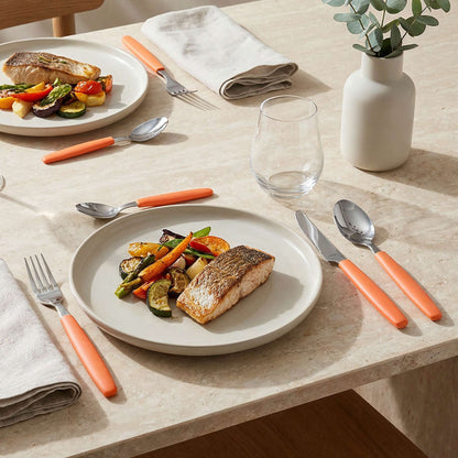 Ein gedeckter Tisch mit einem Teller mit gegrilltem Fisch und gebratenem Gemüse, daneben das 16-teilige Amefa VALOA-Besteckset in Orange, ein Glas, eine helle Serviette und eine weiße Vase mit grünem Laub auf einer hellen Steinplatte.