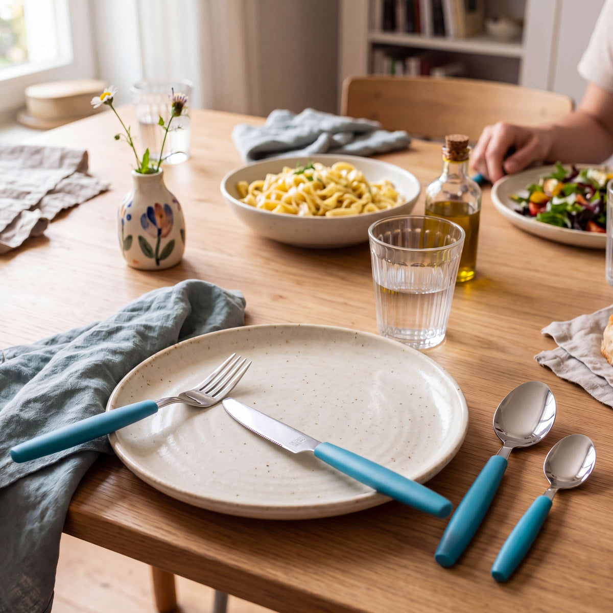Ein Holztisch, gedeckt für eine Mahlzeit mit einem Teller, einer Gabel, einem Messer, einem Löffel, dem 16-teiligen Besteckset „VALOA“ in der Farbe Petrol von Amefa, einem Glas Wasser und einer grauen Serviette. Pasta, Salat, Olivenöl und eine kleine Blumenvase runden das Bild ab.