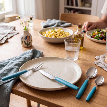 Ein Holztisch, gedeckt für eine Mahlzeit mit einem Teller, einer Gabel, einem Messer, einem Löffel, dem 16-teiligen Besteckset „VALOA“ in der Farbe Petrol von Amefa, einem Glas Wasser und einer grauen Serviette. Pasta, Salat, Olivenöl und eine kleine Blumenvase runden das Bild ab.