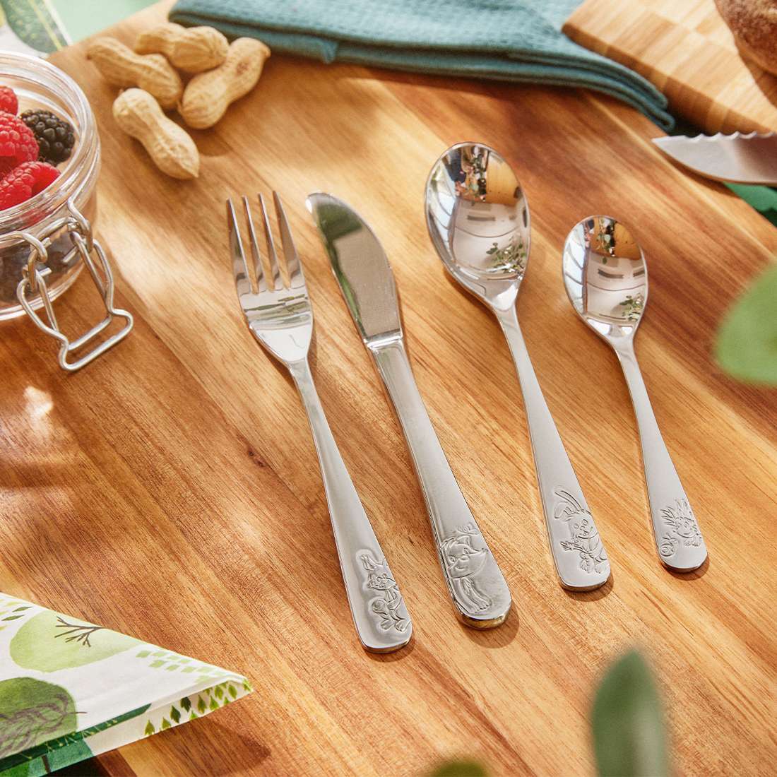 Das Kuppels WALDFREUNDE Kinderbesteck-Set 4-teilig – gravierte Gabel, Messer, großer Löffel und kleiner Löffel aus Edelstahl 18/10 – ist auf einem Holztisch mit Erdnüssen, Beeren im Glas, Servietten und einem blauen Tischtuch arrangiert.