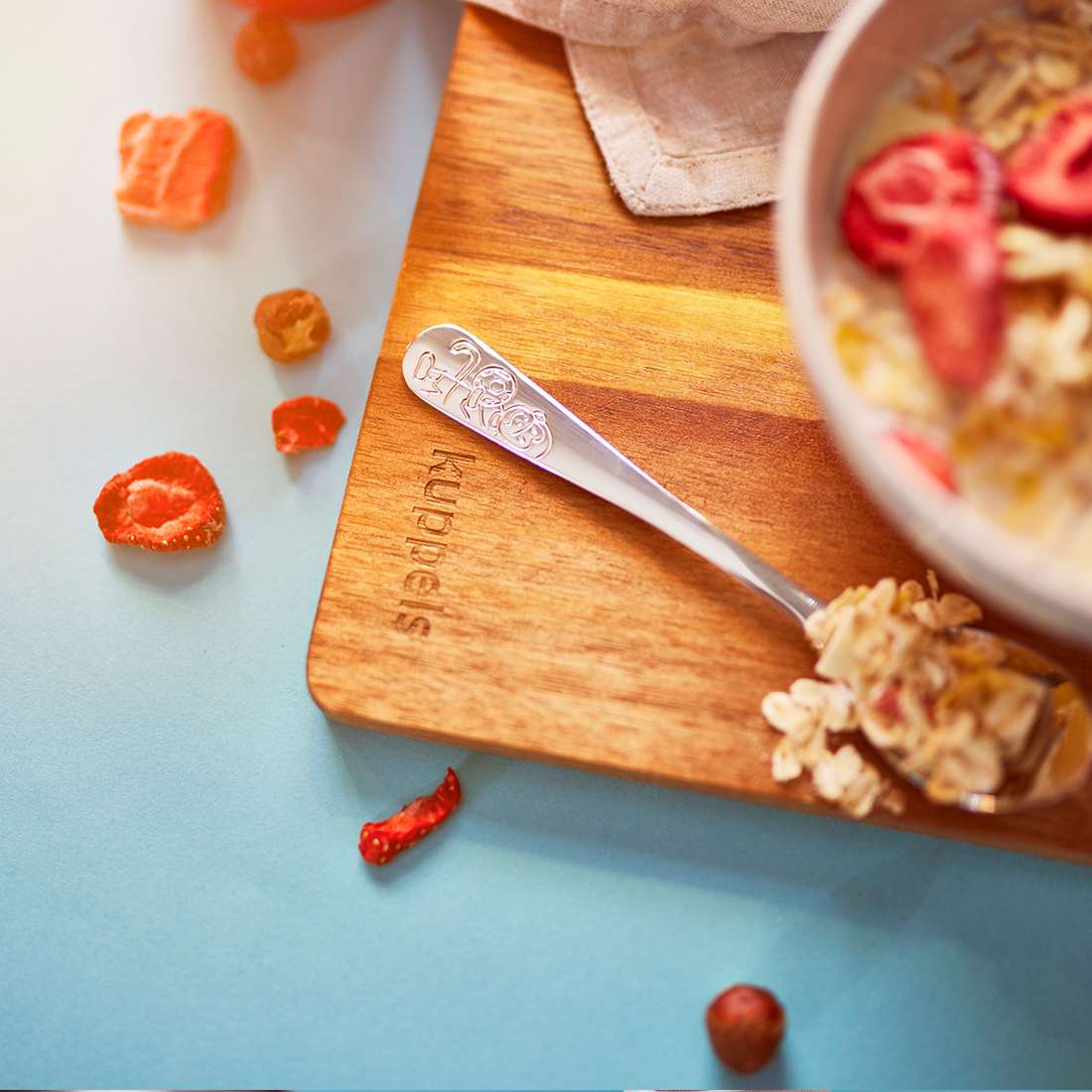 Auf einem Holzbrett steht eine Schüssel Müsli mit Trockenfrüchten. Daneben steht ein Löffel des Kuppels Fußballfreunde Kinderbesteck-Sets 4-teilig (Edelstahl 18/10), auf dem Trockenfrüchte auf einer blauen Fläche verstreut sind.