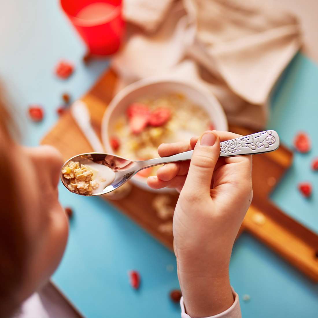 Ein Kind genießt Haferbrei mit Obst mit dem Kuppels Fußballfreunde Kinderbesteck-Set 4-teilig. Die Schüssel steht auf einem Holzbrett mit Obst und einem roten Becher – perfekt für kleine Hände dank des ergonomischen Edelstahl-Designs.