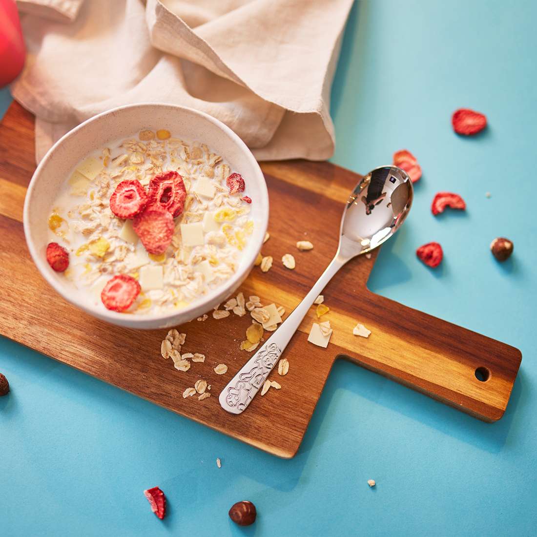 Auf einem Holzbrett steht eine Schüssel Müsli mit Milch, getrockneten Erdbeeren und weißer Schokolade, daneben das 4-teilige Kinderbesteck-Set „Kuppels Fußballfreunde“, vor blauem Hintergrund, auf dem Müsli und Nüsse verstreut sind.