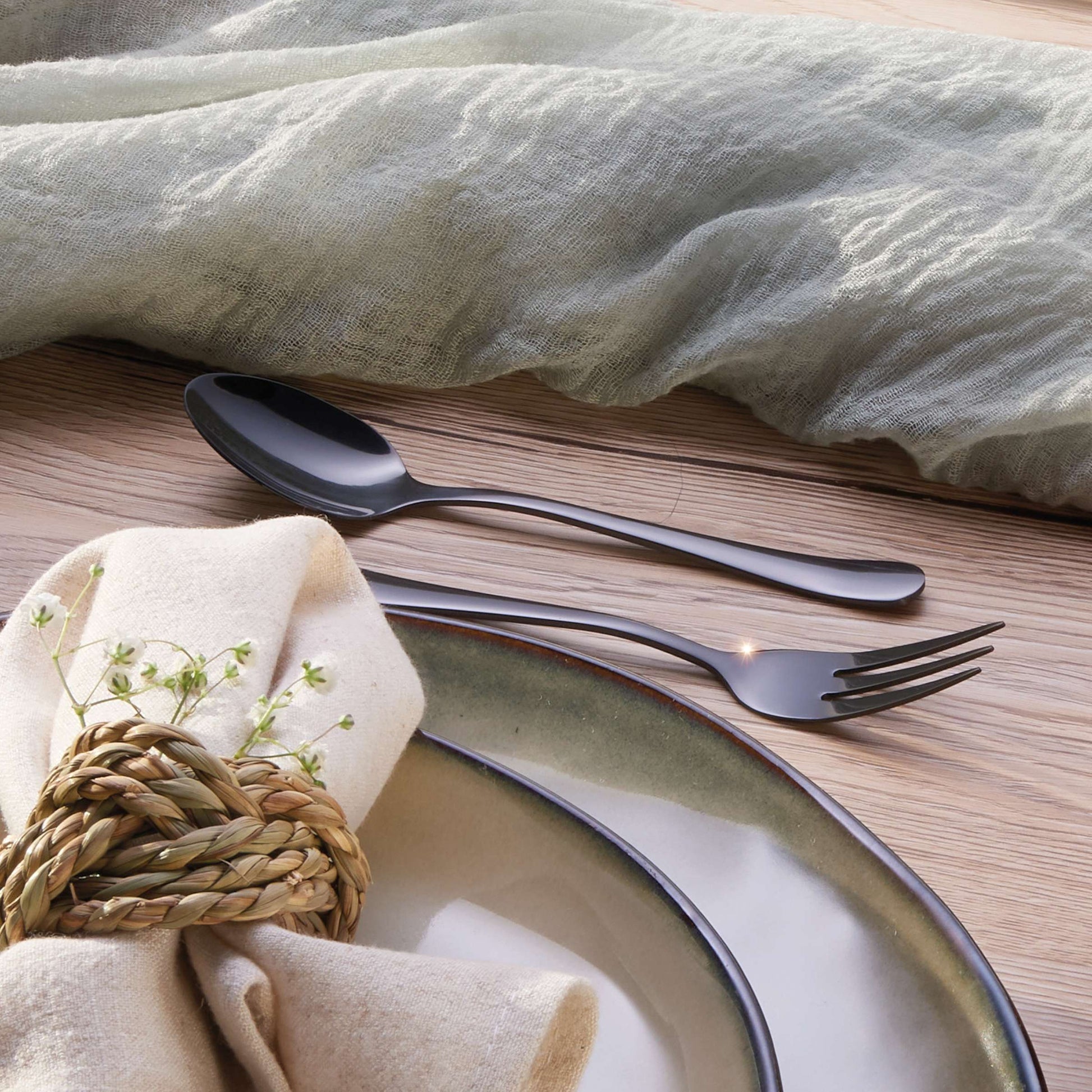 Ein Löffel und eine Gabel aus dem DELIA Besteckset 30-teilig PVD schwarz von Kuppels Home liegen auf einem Holztisch neben zwei übereinander gestapelten Tellern, einer Stoffserviette mit gewebtem Ring und einer hellgrünen Gaze-Tischdecke.