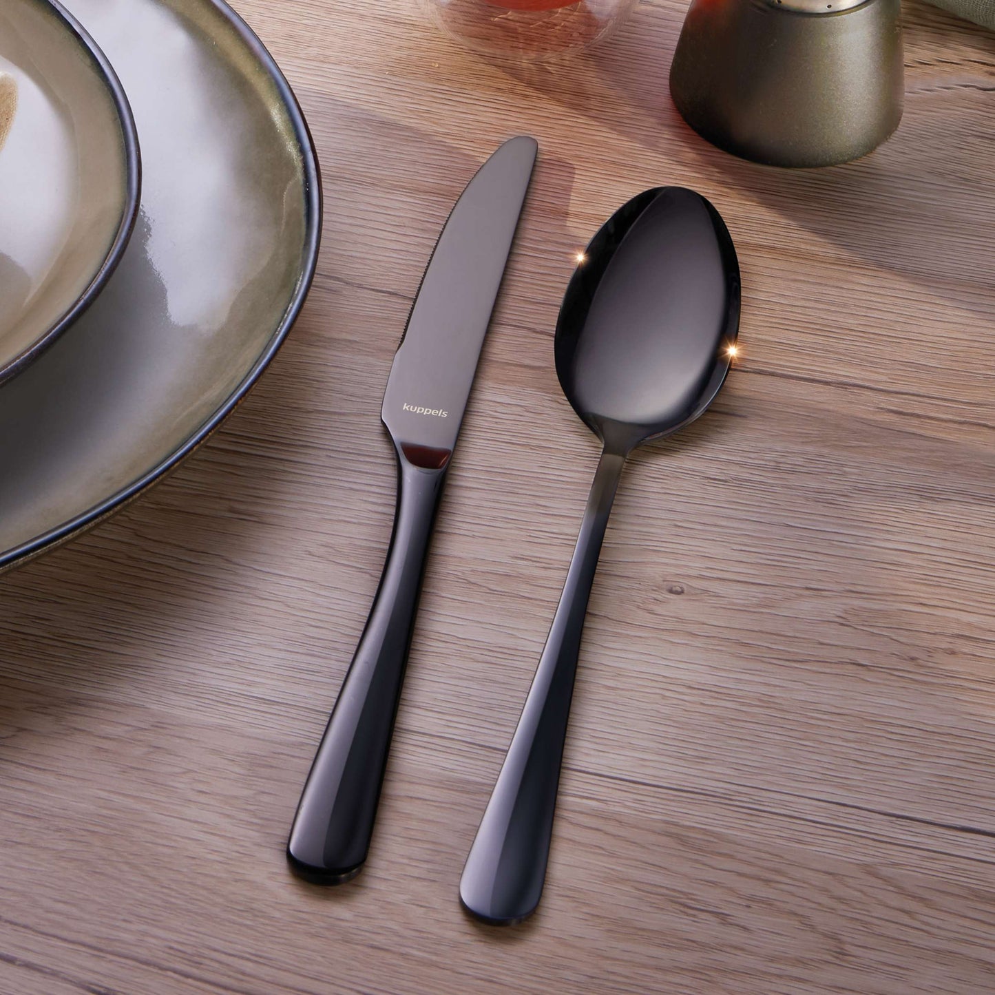 Ein Messer und ein Löffel aus dem „Kuppels Home DELIA Besteckset 30-teilig PVD schwarz“ liegen nebeneinander auf einem Holztisch neben einem Keramikteller mit metallischem Rand.