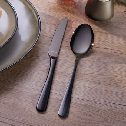 Ein Messer und ein Löffel aus dem „Kuppels Home DELIA Besteckset 30-teilig PVD schwarz“ liegen nebeneinander auf einem Holztisch neben einem Keramikteller mit metallischem Rand.
