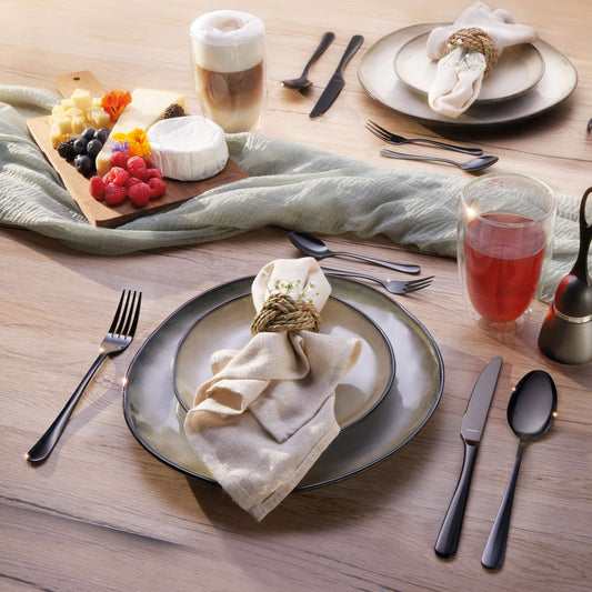 Ein Holztisch, gedeckt mit Tellern, beigen Servietten und dem eleganten 30-teiligen Besteckset „DELIA“ in PVD-Schwarz von Kuppels Home, dazu eine Käse- und Obstplatte, ein Latte und ein Glas Saft. Unter dem Geschirr liegt eine hellgrüne Tischdecke.
