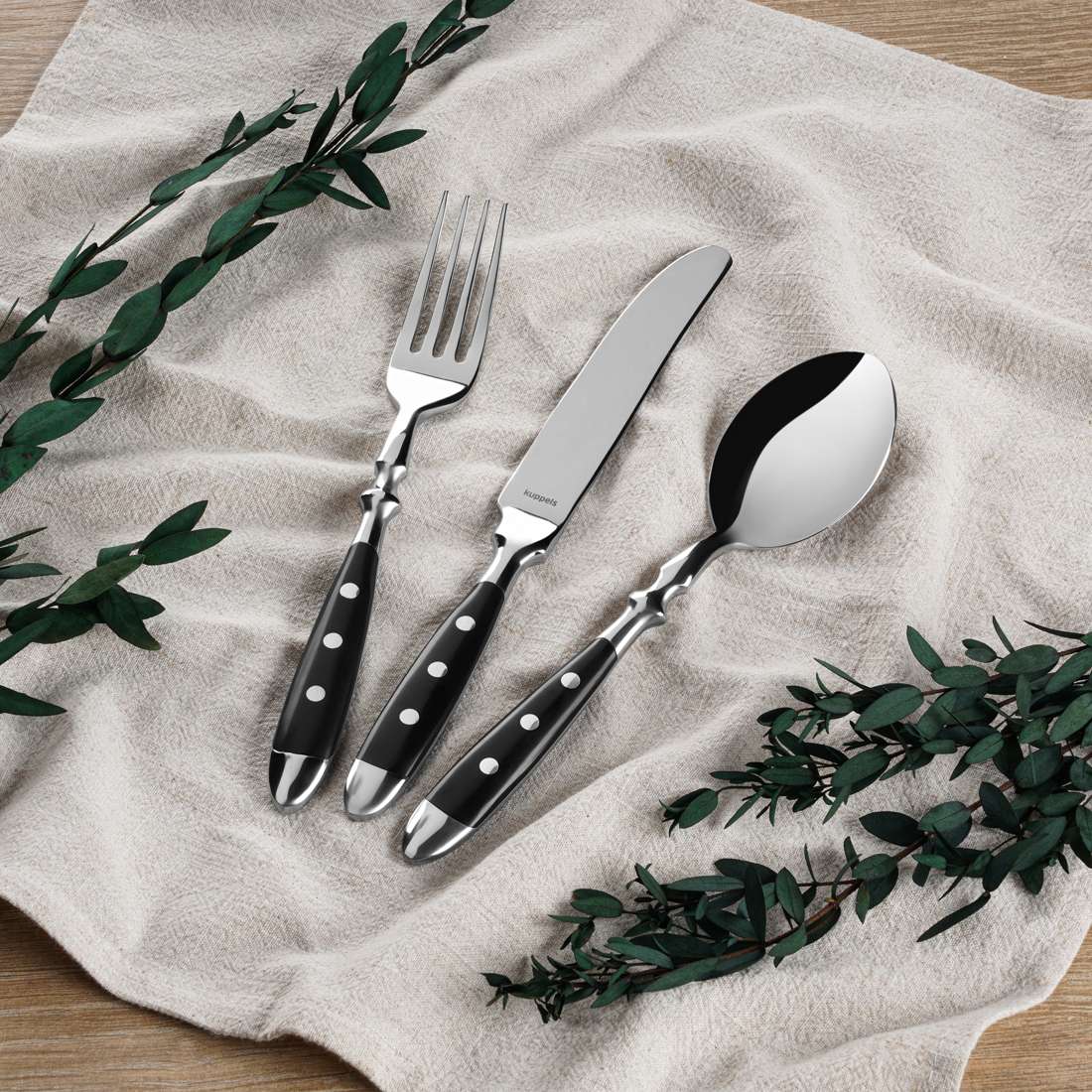 Gabel, Messer und Löffel aus dem Kuppels COUNTRY Besteckset 24-teilig schwarz, mit schwarzen Griffen und silbernen Punkten, sind auf einem beigen Tuch mit grünen Blättern auf einer Holzfläche im gemütlichen Landhausstil angeordnet.
