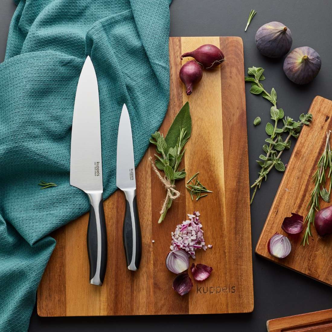 Zwei Kuppels CHEF Tranchiermesser mit Edelstahlklingen liegen auf einem Holzbrett neben einem blaugrünen Tuch, umgeben von gehackten und ganzen roten Zwiebeln, Kräutern und Feigen – ideal für eine elegante Kaffeetafel.