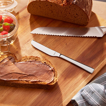 Auf einem Holzschneidebrett liegen eine Scheibe Brot mit Schokoladenaufstrich, ein Glas Beeren, ein Brotlaib, ein gezacktes Brotmesser und ein elegantes Kuppels Edelstahl-Buttermesser aus dem 2-teiligen SELECTION-Buttermesser-Set.