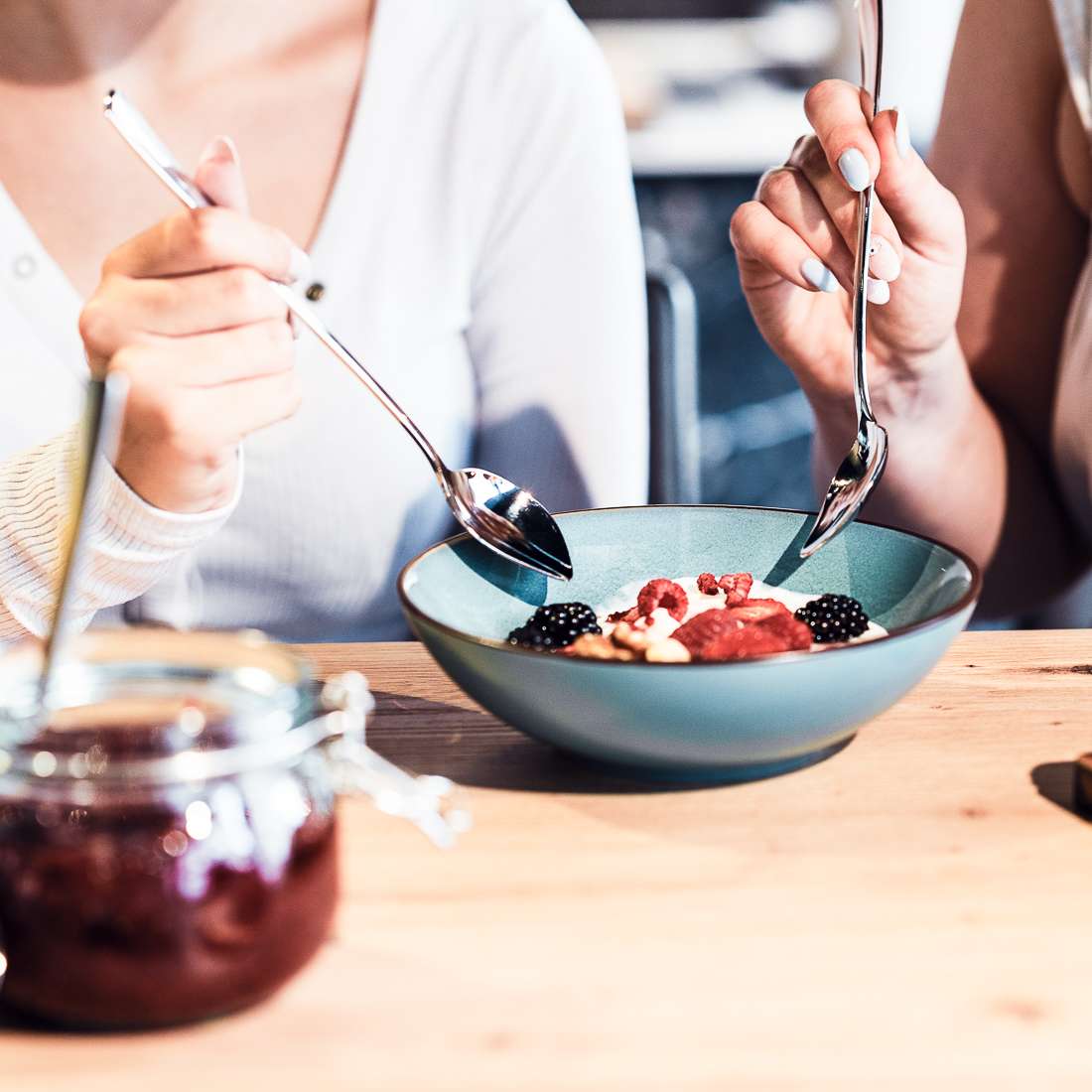 Zwei Personen sitzen mit je einem Kuppels SELECTION Joghurtlöffel aus dem 2er-Set an einem Tisch und teilen sich Joghurt mit Beeren und Granola. Im Vordergrund glänzt ein eleganter Löffel aus Edelstahl 18/10 neben einem Marmeladenglas.