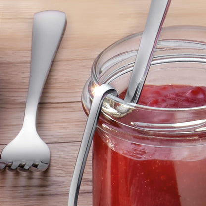 In einem mit roter Marmelade gefüllten Gläschen befindet sich ein Marmeladenlöffel der Marke Kuppels SELECTION, während am Rand eine Gabel mit hakenförmigem Griff befestigt ist und ein weiteres Besteckteil auf dem Holztisch liegt.