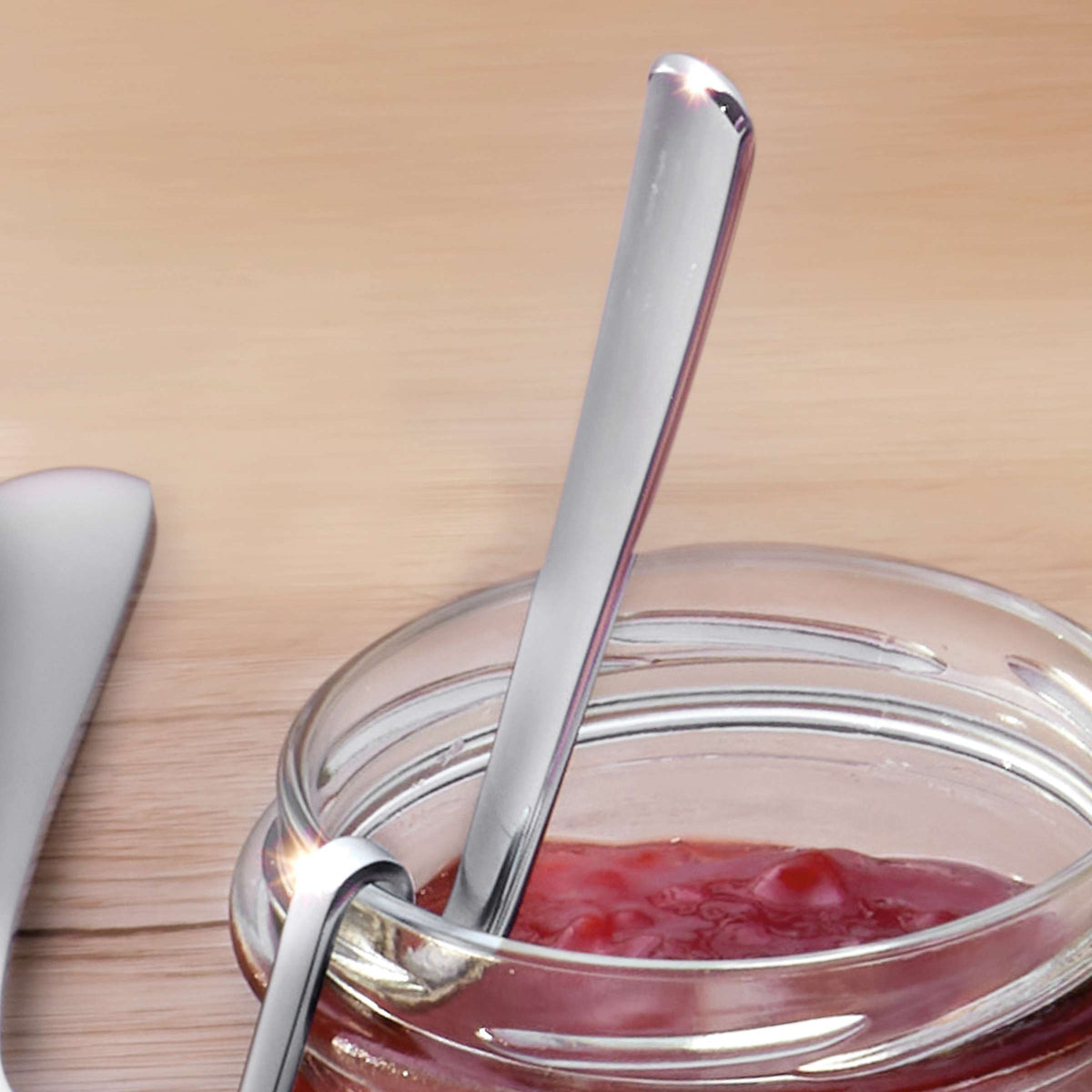 Eine Auswahl an Marmeladenlöffeln von Kuppels liegt in einem mit roter Marmelade gefüllten Glas, das auf einer Holzunterlage steht, auf der links ein weiteres Besteckteil zu sehen ist.