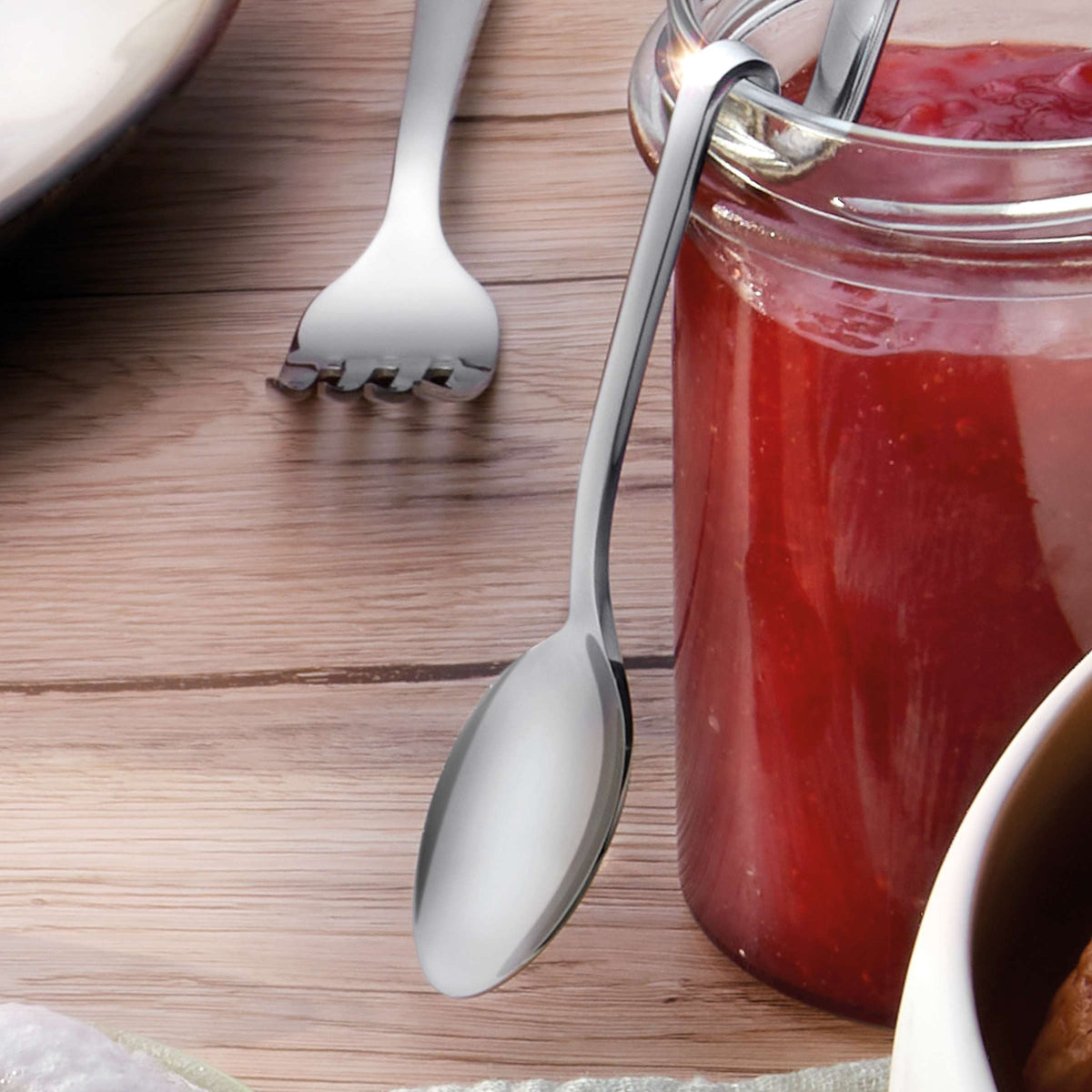 Eine Auswahl an Marmeladenlöffeln von Kuppels liegt auf einem Holztisch neben einem Glase mit Erdbeermarmelade; im Hintergrund sind ein Teil einer Gabel und ein Teller zu sehen.
