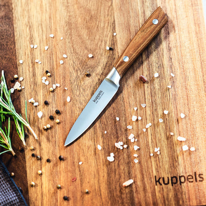 Ein AVID-Gemüsemesser von Kuppels mit kompaktem Design und einem Griff aus Eschenholz liegt auf einem Holzschneidebrett, das mit Salz, Pfeffer und Kräutern bestreut ist; auf dem Brett ist das Wort „kuppels“ eingebrannt.