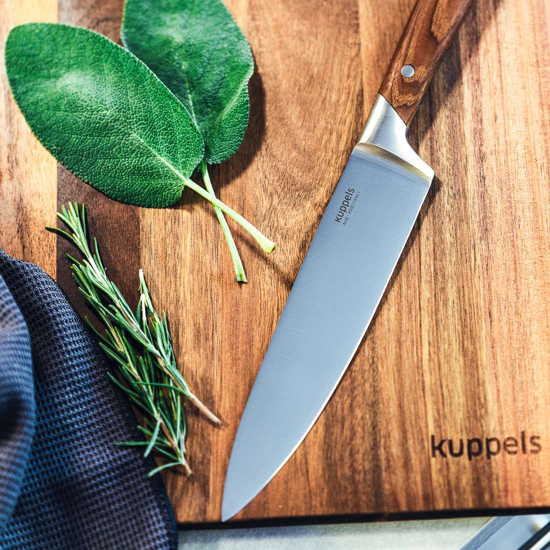 Ein AVID Kochmesser 8" von Kuppels mit Eschenholzgriff liegt auf einem hölzernen Schneidebrett von Kuppels neben frischem Rosmarin, Salbeiblättern und einem dunklen Tuch.