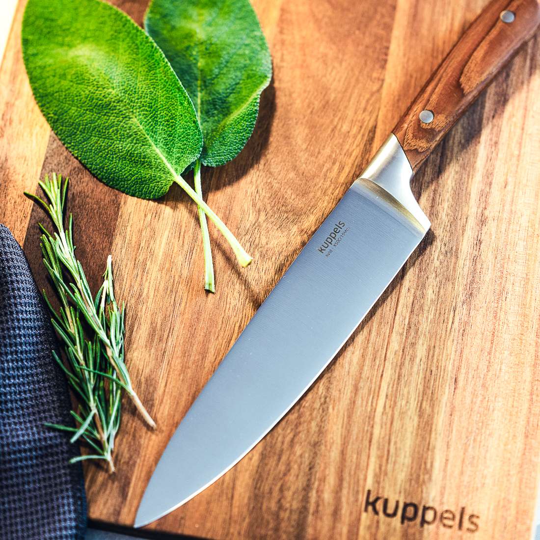 Ein AVID Kochmesser 8" von Kuppels mit Eschenholzgriff liegt neben zwei Salbeiblättern und Rosmarinzweigen auf einem Holzschneidebrett. Der Markenname von Kuppels ist in die Ecke des Bretts eingraviert.