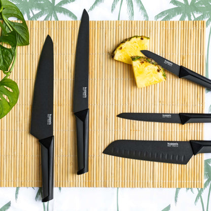 Fünf schwarze Santokumesser der Marke „Kuppels BLACK“ mit scharfen Klingen und schwarzen Griffen liegen auf einer Bambusmatte neben zwei Ananasstücken. Im Hintergrund ist ein weißes Tuch mit grünen Palmblattmustern zu sehen, und oben links befindet sich ein grüner Zweig.