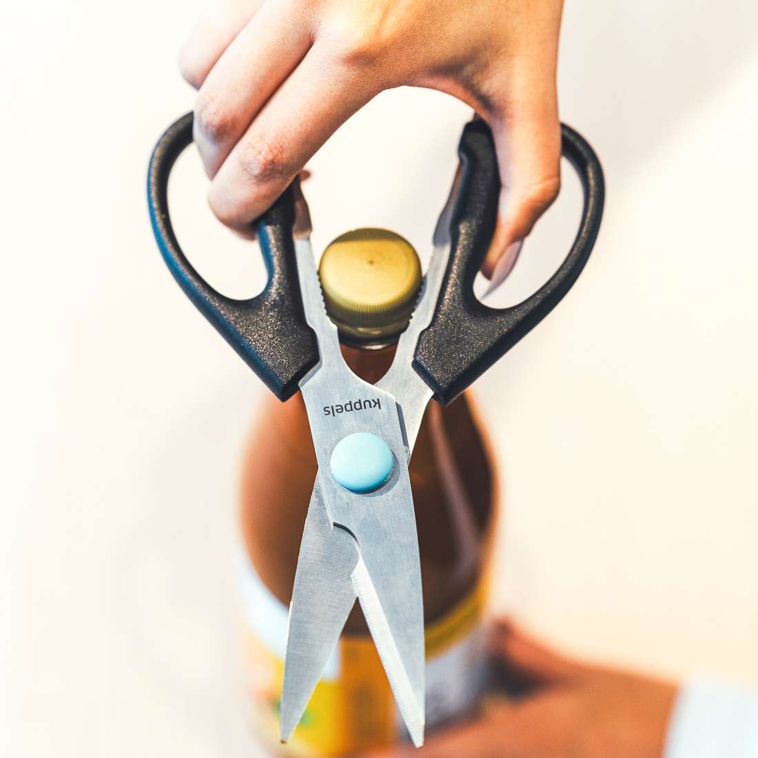 Eine Hand verwendet die Kuppels SELECTION Allzweckschere mit scharfen Klingen und Komfortgriffen, um eine Flasche mit einem goldenen Verschluss zu greifen und aufzudrehen, wobei sie die Schere um den Verschluss legt und so ein müheloses Öffnen ermöglicht.