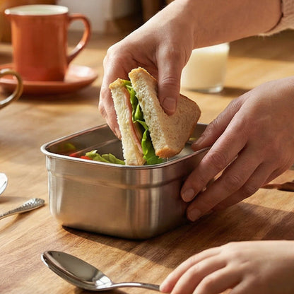 Eine Person legt ein Sandwich mit Salat in eine „Kuppels SELECTION“ Frischhaltedose (2-teilig) auf einem Holztisch ab; daneben stehen ein Löffel, eine Gabel, eine Tasse und ein Glas Milch.